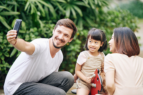 Cheerful young Caucasian man taking selfie with his pretty Vietnamese wife and daughter at playground