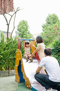 Happy young parents helping their daughter to slide down at playground