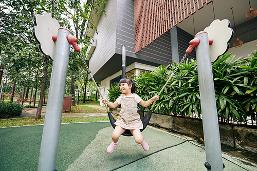 Cheerful excited little Asian girl enjoying swinging on playground of kidergardern