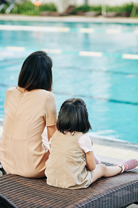 Mother and little daughter sitting on chaise-lounge and looking at hotel swimming pool, view from the back