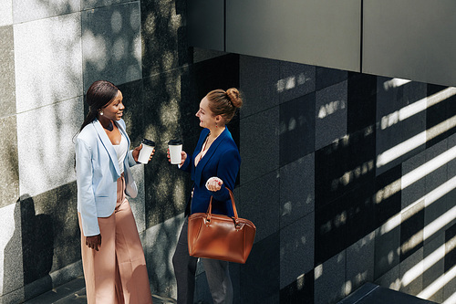 Smiling businesswomen standing on stairs outdoors with take out coffee and gossiping about work
