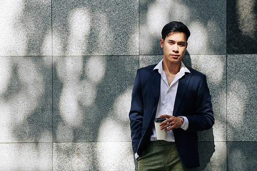Portrait of young attractive Asian man standing at building wall and drinking cup of coffee