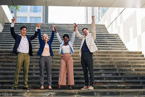 Excited happy young business people holding hands and raising arms when standing on steps outdoors