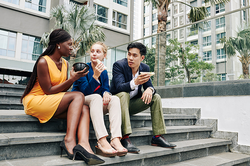 Multi-ethnic colleagues sitting on steps outdoors, eating lunch and gossiping