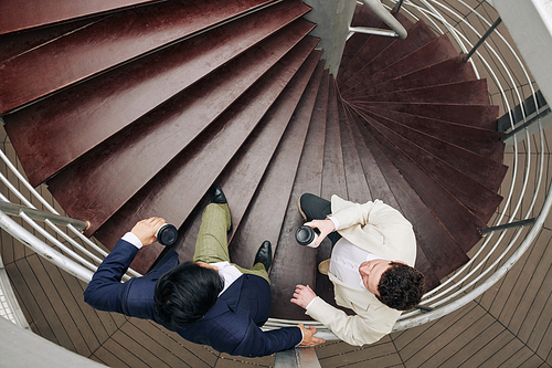 Young business people standing on spiral staircase, drinking take out coffee and discussing gossips and work details, view from above