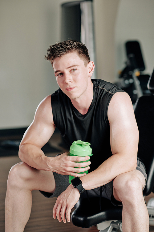 Portrait of handsome young smiling man drinking protein shake when sitting on bench in gym