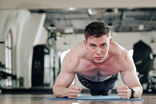 Determined shirtless young man standing in plank position on yoga mat in local gym