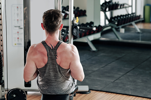 Fit strong man doing exercise on rowing machine in sports center, view from the back