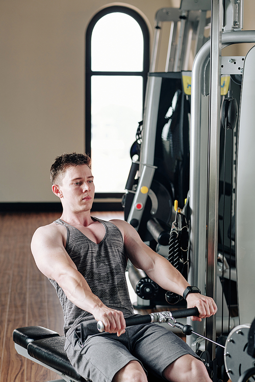 Serious handsome young bodybuilder concentrated on doing exercise in rowing machine in gym