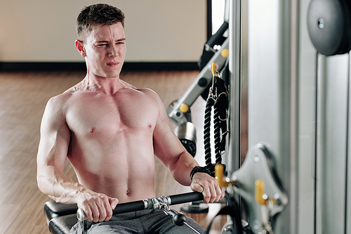 Shirtless fit young man pulling cable of rowing machine when having functional training in gym