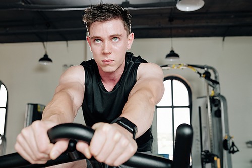 Serious handsome young sportsman riding stationary bicycle when having cardio training in gym