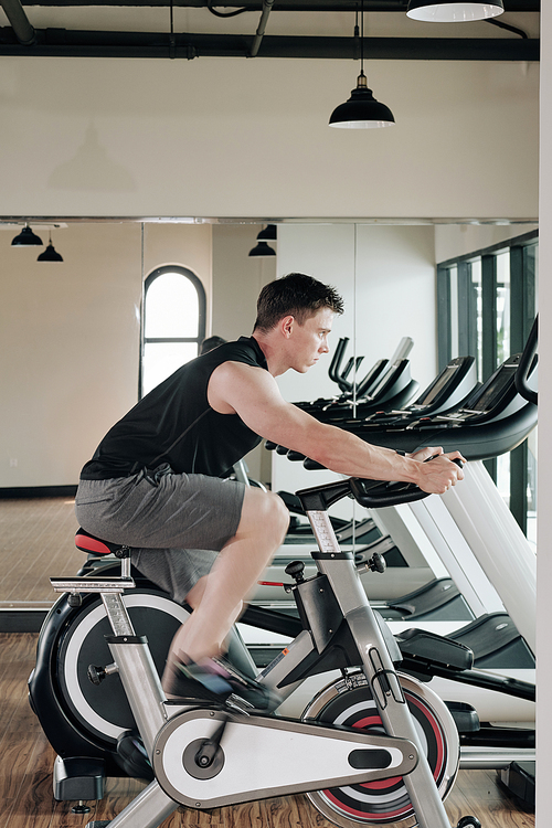 Serious sportsman training at fitness center and riding stationary bike at high speed