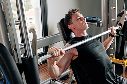 Muscular young man sitting on gym bench and lifting heavy barbell