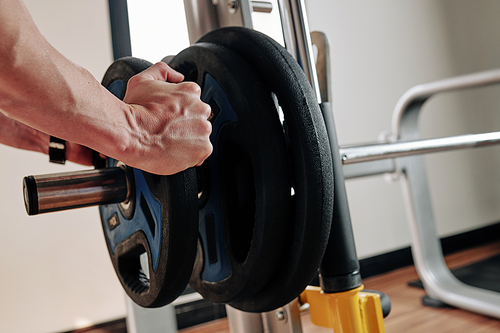 Close-up image of sportsman putting heavy weight plates on barbell before exercising in gym