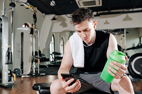 Fit young man setting music on his smartphone and drinking vitamin water between sets