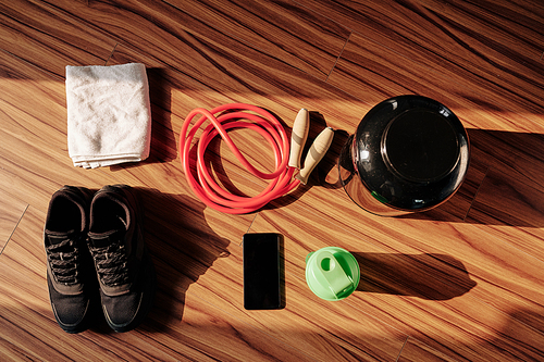 Belongings of sportsman on wooden floor: sneakers, skipping rope, shaker with protein cocktail, smartphone and towel