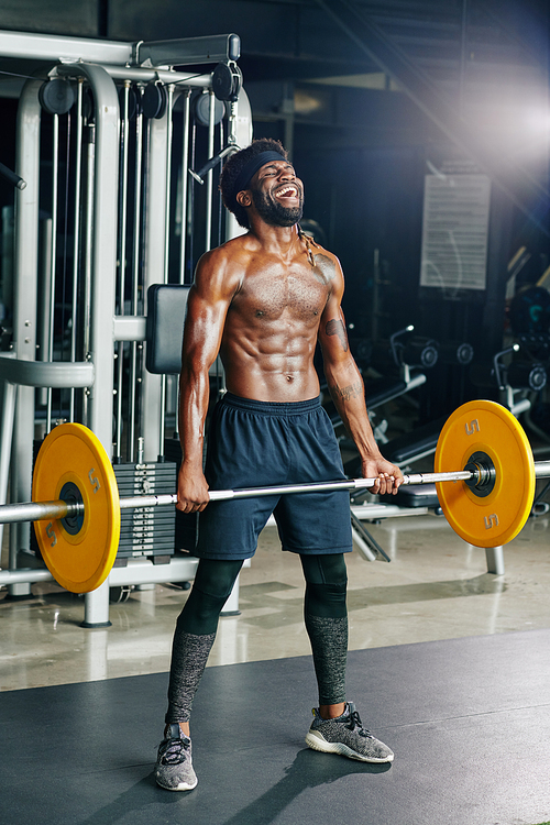 Vertical full shot of handsome African man with defined body standing shirtless holding barbell