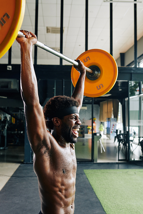 Vertical medium shot of strong African man standing shirtless doing lifting up barbell exercise in gym