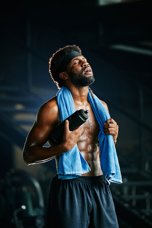 Vertical portrait of tired muscular African man in sweat with towel on shoulders having break during workout