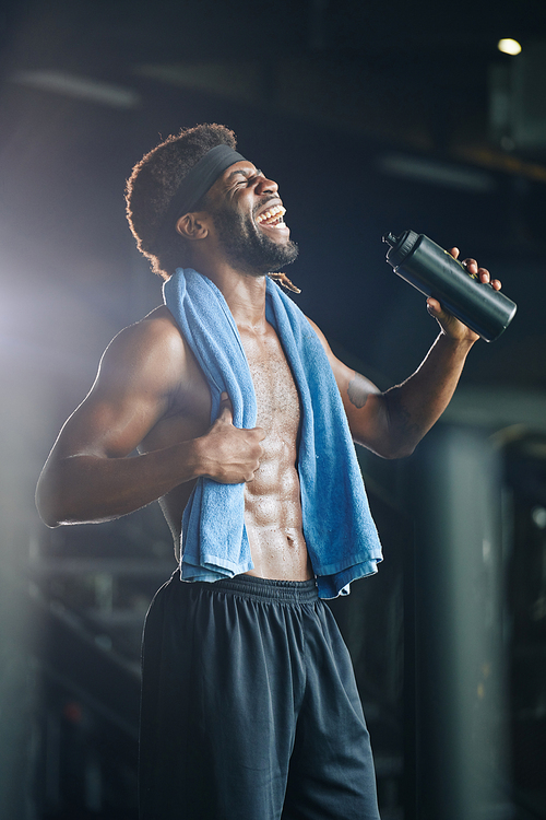 Vertical medium long shot of cheerful masculine guy standing topless with towel on shoulders holding sport bottle with water