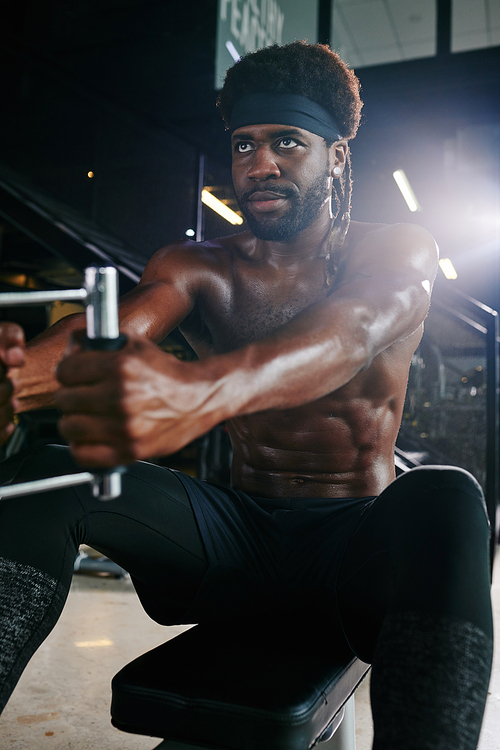 Vertical portrait of sporty African American man doing strengthening exercise in gym using fitness machine