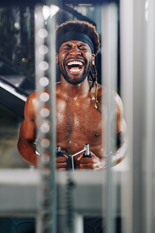 Vertical portrait of African American man laughing at something while doing exercise using fitness machine in gym