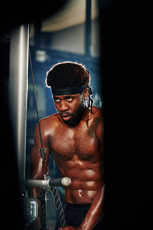 Vertical portrait of concentrated African American man exercising in modern gym using fitness equipment