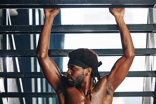 Horizontal shot of strong black man practicing pull-up exercise in modern gym looking away