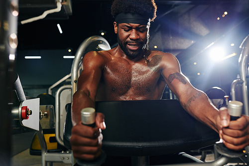 Horizontal shot of masculine black guy doing strength exercise using gym equipment