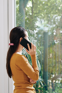 Young woman in mustard color sweater looking through window when calling on phone