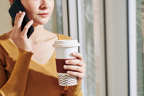 Cropped image of serious young businesswoman drinking big cup of take out coffee and talking on phone