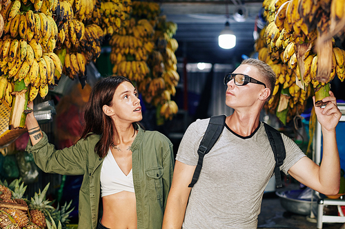 Young man and woman walking along Asian farmer's market looking at bananas and chatting, horizontal shot