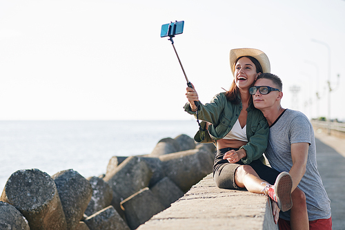 Horizontal shot of happy young man and woman spending time together at seaside taking selfie on phone using selfie stick