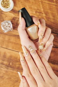 Hands of woman with fancy manicure with rhinestones holding bottle of golden nail polish