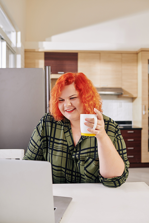 Smiling overweight young woman with red hair drinking morning coffee in kitchen and checking e-mail on her laptop