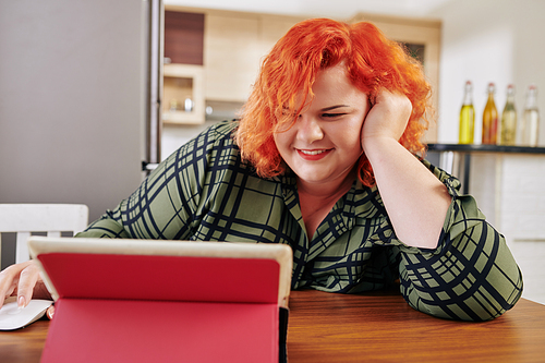 Cheerful overweight woman enjoying watching captivating movie on tablet computer at home