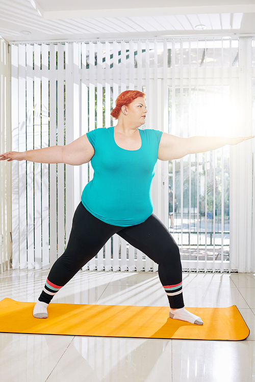 Plus size young woman standing in warrior pose when praciting asana on yoga mat at home
