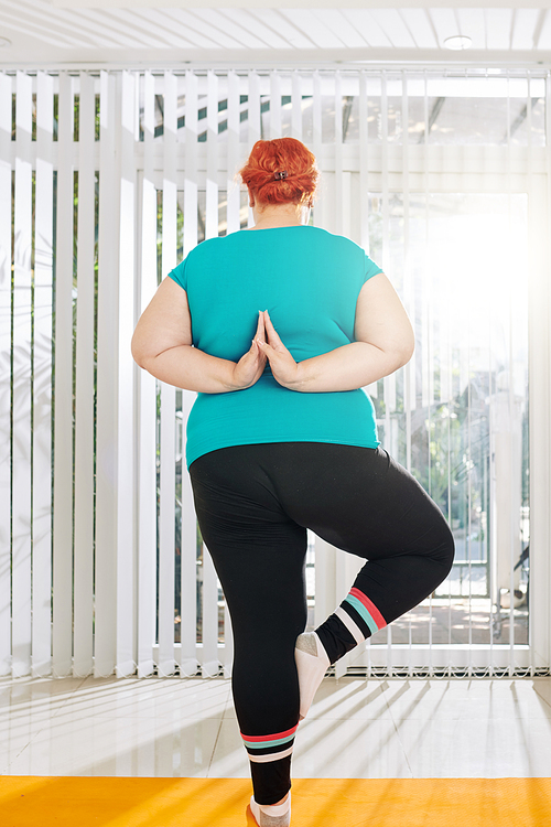 Overweight woman standing on one leg and keeping hands in reverse namaste mudra behind the back