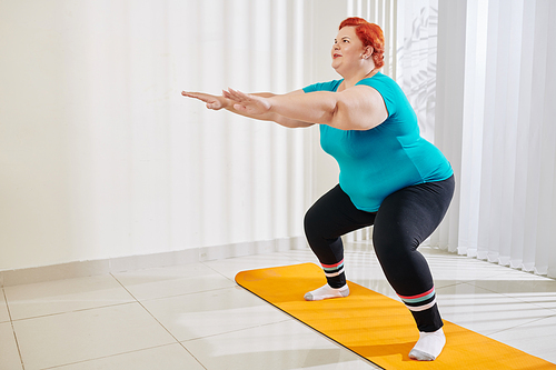 Smiling overweight woman outstretching arms for balance when doing squats at fitness class