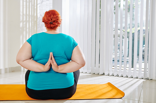 Rear view of overweight woman sitting on yoga mat and keeping hands in reverse namaste mudra behind her back