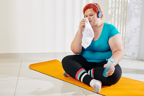 Young overweight woman wiping face and drinking fresh water after training at health club