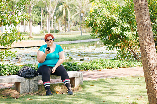 Overweight young woman in sunglasses resting on bench in park and reading text messages on smartphone