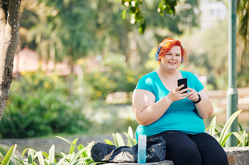 Portrait of young smiling plus size woman sitting on bench in park and checking application in her smartphone