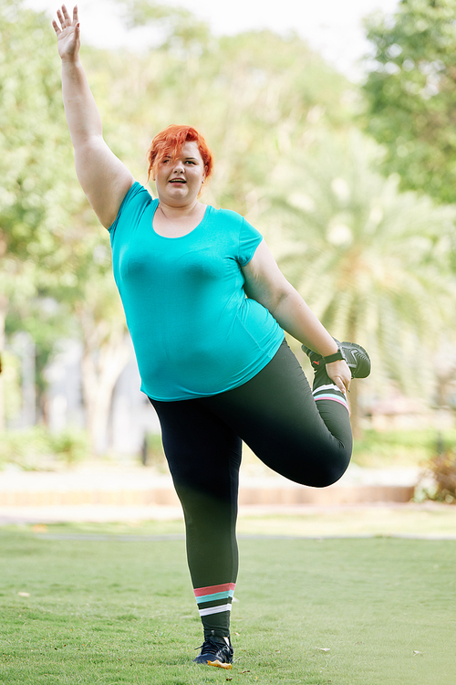 Overweight young woman standing on one leg and raising opposite arm when doing balance exercise in park