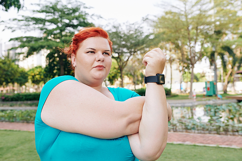 Serious plus size young woman doing stretching exercise for her arms when warming up in park
