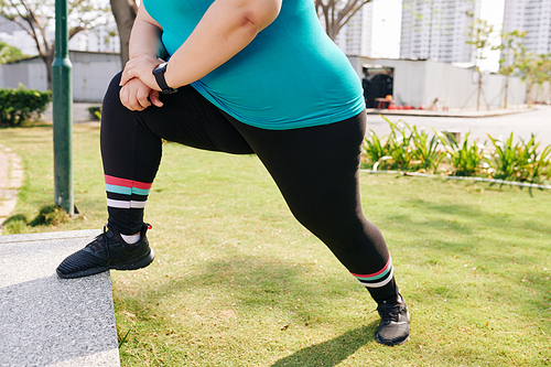 Cropped image of young overweight woman putting leg on parapet and stretching it after jogging