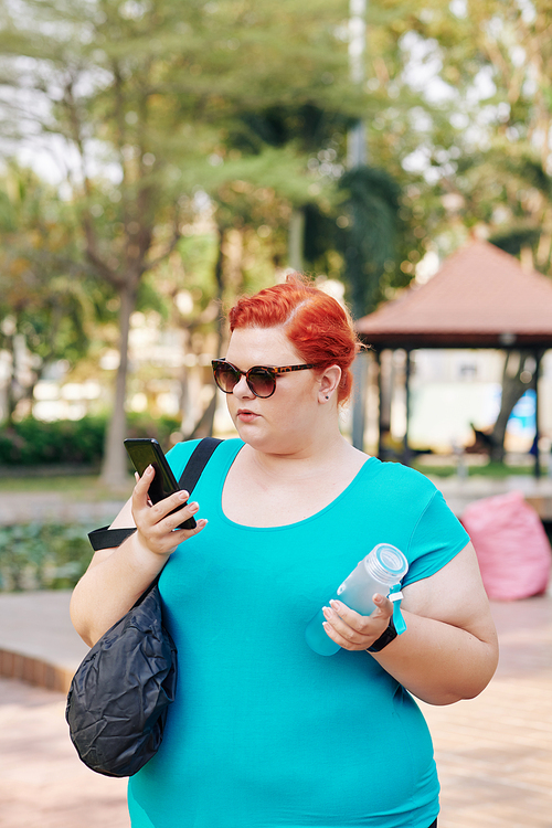 Plus size woman in sunglasses walking outdoors and reading text messages from her friends