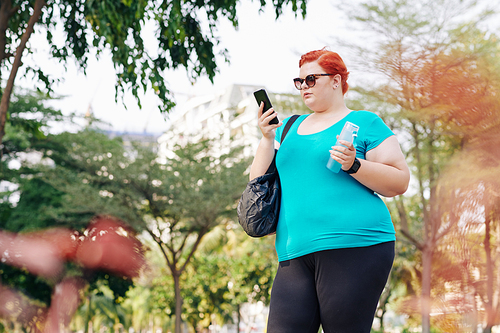 Overweight pretty young woman standing in city park and checking health application on smatphone after training