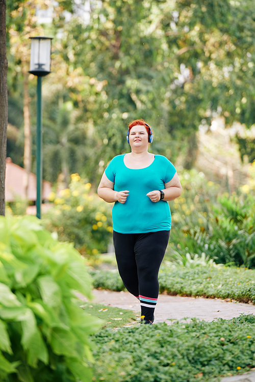 Smiling overweight young woman in headphones jogging in city park