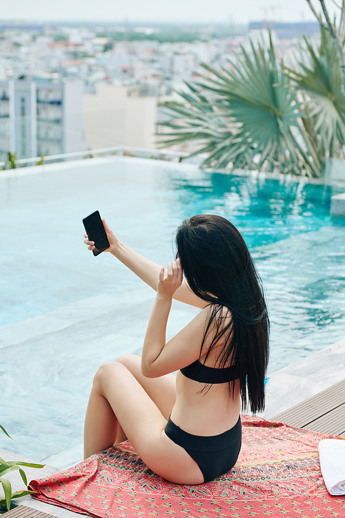 Pretty young woman in bikini taking selfies when sitting by swimming pool
